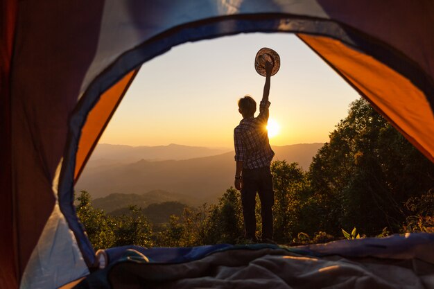 幸せな男は、レジャーと自由を楽しんで夕焼けの光の空の下で山の周りのテントの近くに滞在します。