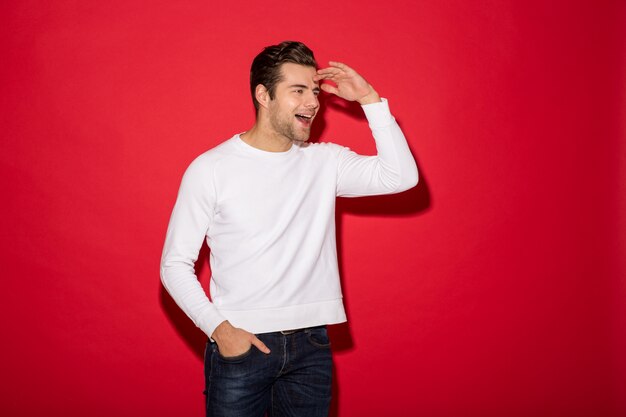 Happy man in sweater looking away with open mouth and arm in pocket over red wall