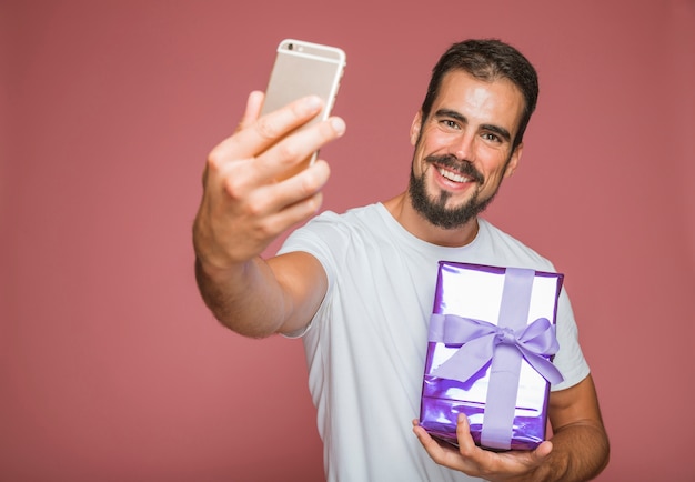 Foto gratuita uomo felice che prende selfie con il cellulare che tiene il contenitore di regalo