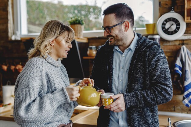 Happy man talking to his wife while she is pouring him a cup of tea in the kitchen