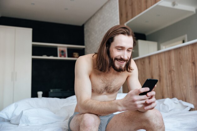 Happy man using cell phone sitting on bed in bedroom