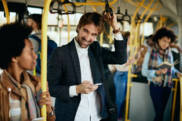 Счастливый человек пользуется мобильным телефоном и разговаривает с пассажиром во время поездки на автобусе