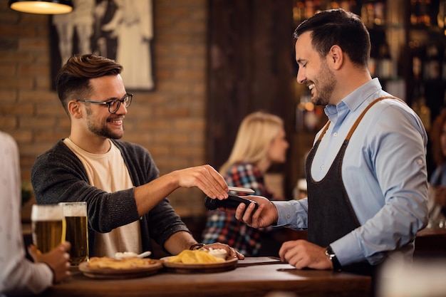 Happy man using smart phone and paying for the bill via contactless payment in a pub