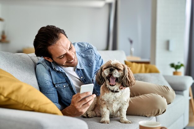 Happy man using smart phone while relaxing on the sofa with his dog who is yawning