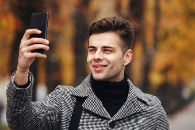 Happy man warmly dressed taking photo of nature or making selfie using black smartphone, while walking in park