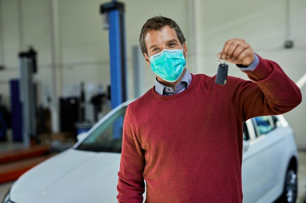 Happy man wearing protective face mask while holding keys of his repaired car in a workshop