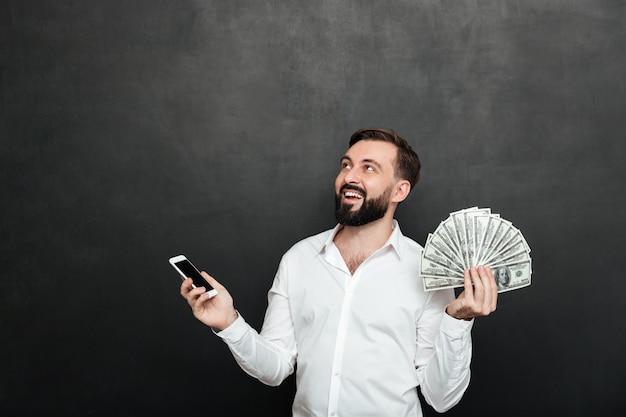 흰 셔츠에 행복 한 사람이 그의 휴대 전화를 사용 하여 현금 돈을 많이 승리 하 고 어두운 회색을 올려
