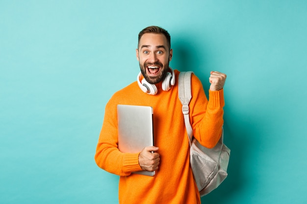 Happy man with backpack and headphones, holding laptop and smiling, cheering of win, triumphing
