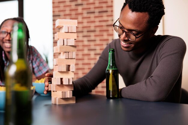 Happy man with beverage on table carefully removing wooden block from wood tower. Smiling person sitting at home in living room with close friends while playing society games together.