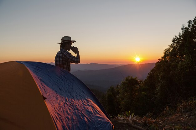 ドリンクを飲みながら幸せな男は夕日の光の下で山の周りのテントの近くに滞在します。