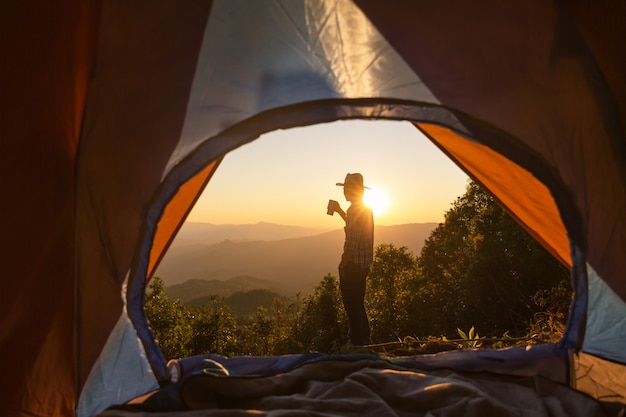 コーヒーカップを持って幸せな男は山の周りのテントの近くに滞在します。