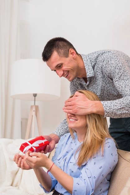 Happy man with present closing eyes to smiling woman on sofa