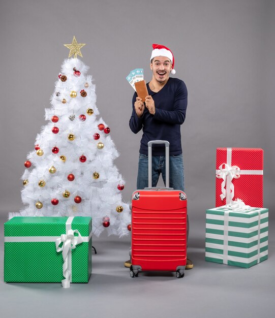 happy man with red suitcase showing his travel tickets on grey