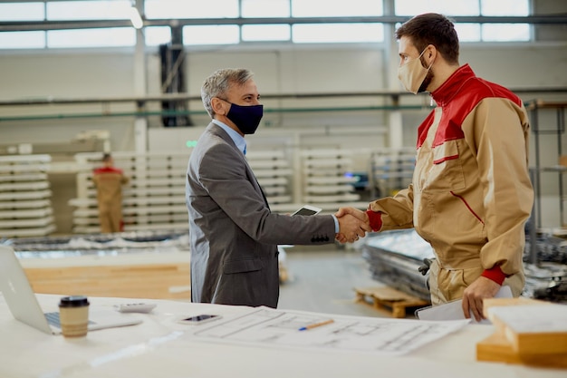 Happy manager and manual worker wearing face masks while handshaking at carpentry workshop