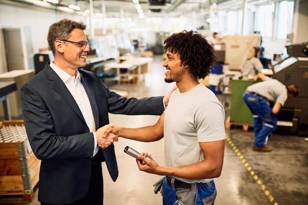 Happy manager shaking hands with African American factory worker in industrial building