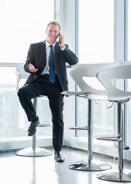 Счастливый зрелый бизнесмен, сидя на стуле, говорить по мобильному телефону