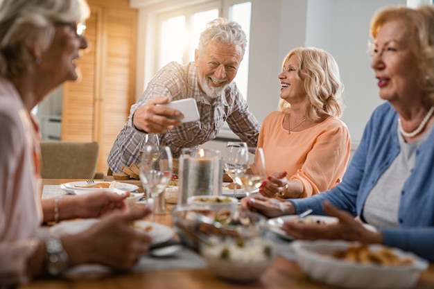 Happy mature couple having fun while having lunch with friends and taking selfie at dining table