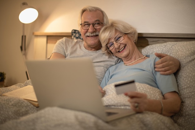 Счастливая зрелая пара развлекается, делая покупки в Интернете с помощью кредитной карты вечером в спальне