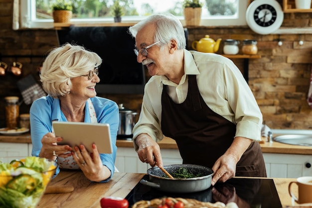 부엌에서 디지털 태블릿을 사용하여 음식을 준비하고 교활한 의사 소통을 하는 행복한 성숙한 부부