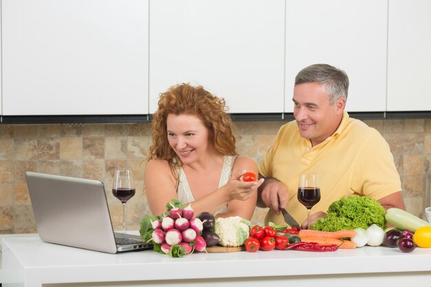 Happy mature couple using laptop computer in home People are in search of recipes for preparing something from fresh vegetables in the kitchen