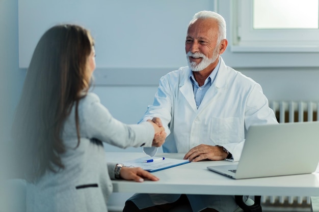 Happy mature doctor and his female patient shaking hands after medical appointment at clinic