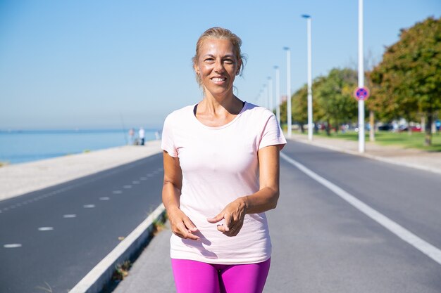 Happy mature female jogger walking down running track at river, looking and pointing finger. Front view. Activity and age concept