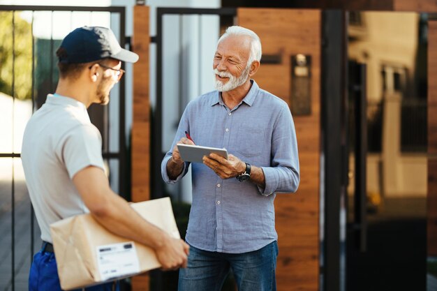 Happy mature man signing on digital tablet for a delivery while communicating with a courier