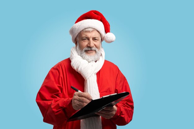 Happy mature Santa Claus with real beard in red hat and white scarf holding pen and black clipboard, isolated on blue background. Senior man planning his day, work and holiday celebration.