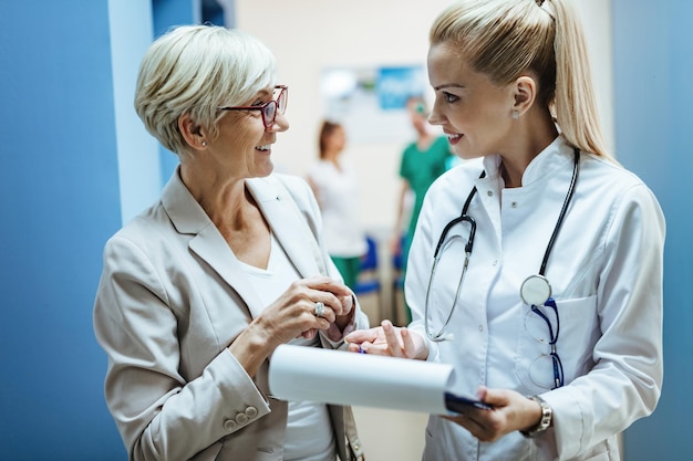 Happy mature woman and her doctor communicating while going through paperwork in a hospital hallway