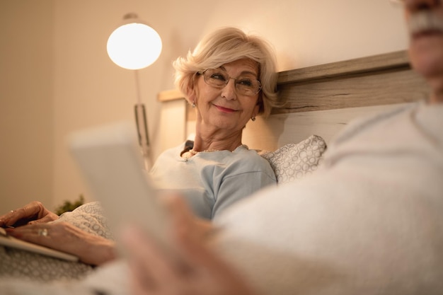 Happy mature woman resting in bed and using laptop while looking at her husband who is lying down next to her