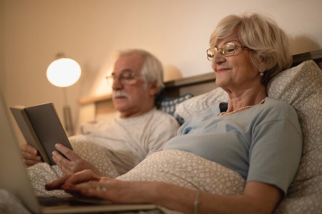 Happy mature woman surfing the net on laptop while resting with her husband in bedroom in the evening