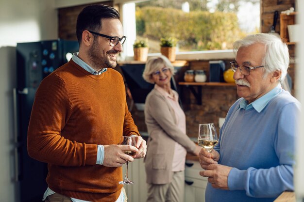 Happy mid adult man and his mature father drinking wine while communicating in the kitchen There is a woman in the background