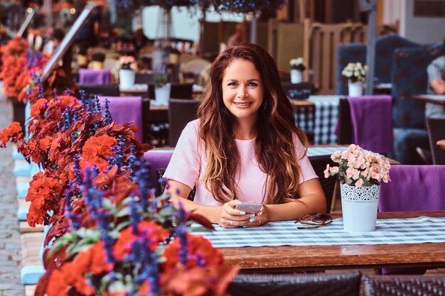 Happy middle age businesswoman with long brown hair holds a smartphone while sitting at an outdoor cafe.