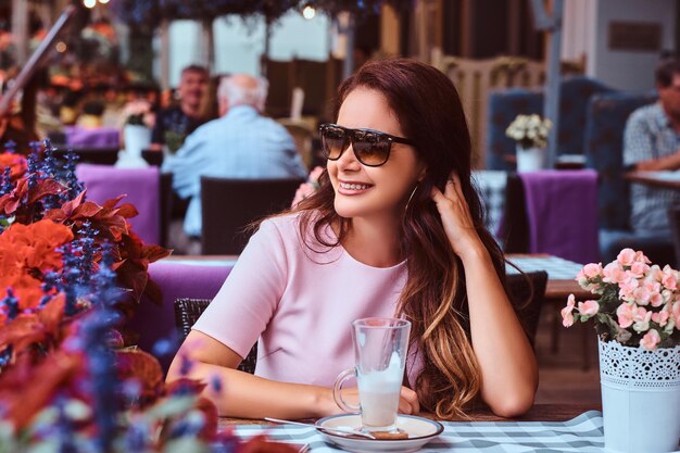 Happy middle age businesswoman with long brown hair wearing a pink dress sitting with a glass of cappuccino at the outdoor cafe.