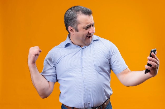 Happy middle age man in blue stripped striped shirt looking at his mobile phone and raising his hand in the gesture of triumph while standing 