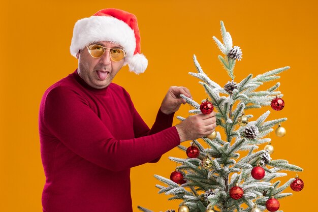 Happy middle age man wearing christmas santa hat in dark red turtleneck and yellow glasses sticking out tongue decorating christmas tree standing over orange wall