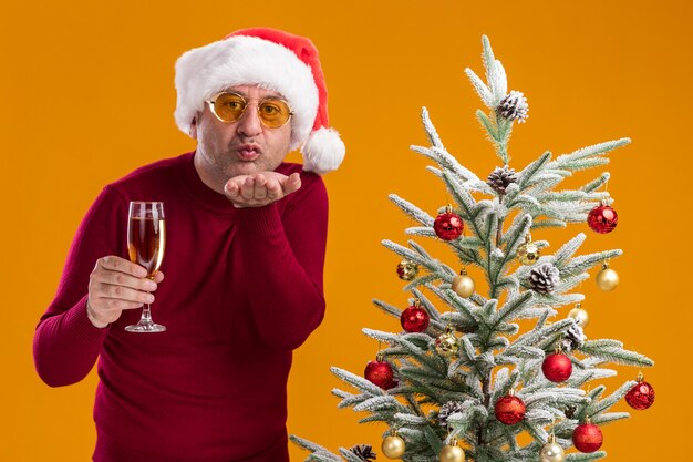 Happy middle age man wearing christmas santa hat in dark red turtleneck and yellow glasses with glass of champagne blowing a kiss standing next to a  christmas tree over orange background