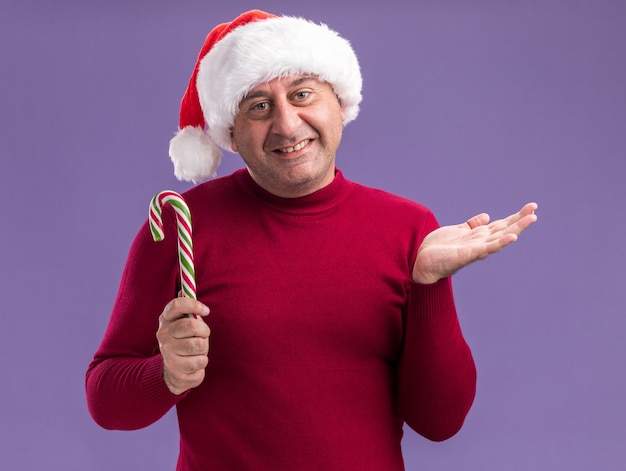 Счастливый мужчина среднего возраста в рождественской шапке санта-клауса, держащий конфету, смотрит в камеру с улыбкой на лице с поднятой рукой, стоя на фиолетовом фоне
