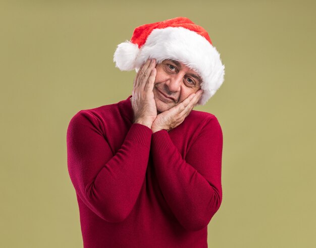 녹색 배경 위에 서있는 얼굴에 미소로 카메라를보고 크리스마스 산타 모자를 쓰고 행복한 중년 남자