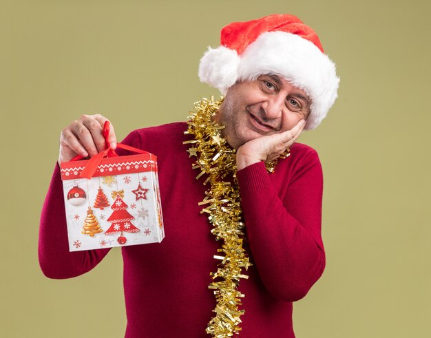 happy middle age man wearing christmas santa hat with tinsel around neck holding christmas gift looking at camera smiling cheerfully standing over green background