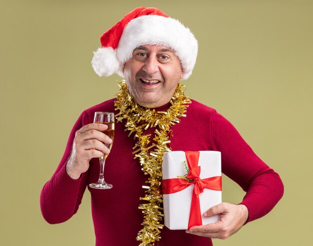 幸せな中年の男は、クリスマスプレゼントと緑の背景の上に立っている顔に笑顔でカメラを見てシャンパンのガラスを保持している首の周りに見掛け倒しのクリスマスサンタの帽子をかぶっています