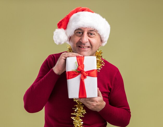 Happy middle age man wearing christmas santa hat with tinsel around neck holding christmas  present  with smile on face  standing over green  wall