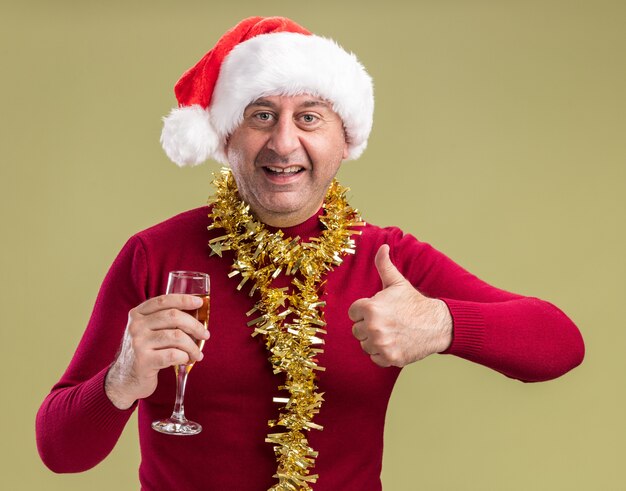 首の周りに見掛け倒しのクリスマスサンタの帽子をかぶって幸せな中年の男は、緑の背景の上に立って親指を見せて笑っているカメラを見てシャンパンのガラスを保持しています