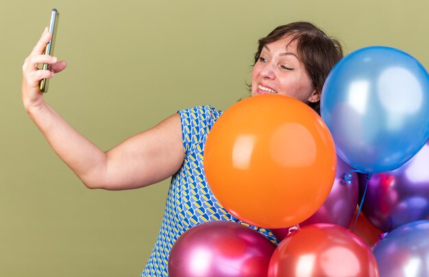 Happy middle age woman bunch of colorful balloons doing selfie using smartphone smiling cheerfully