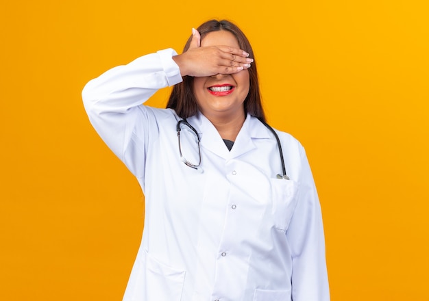 オレンジ色の壁の上に立っている手で目を覆う聴診器と白衣で幸せな中年女性医師 Premium写真
