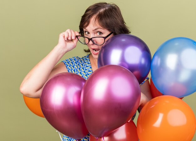 Happy middle age woman in glasses with bunch of colorful balloons  surprised