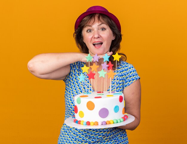 Happy middle age woman in party hat holding birthday cake  smiling cheerfully