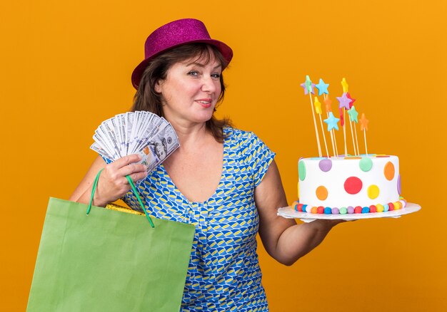 Happy middle age woman in party hat holding paper bag with gifts holding birthday cake and cash  smiling broadly celebrating birthday party standing over orange wall