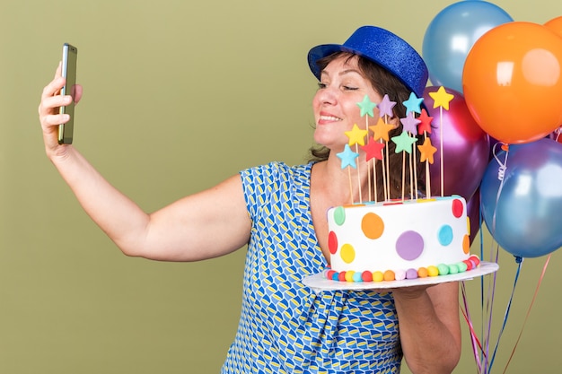 Happy middle age woman in party hat with bunch of colorful balloons holding birthday cake doing selfie using smartphone celebrating birthday party standing over green wall