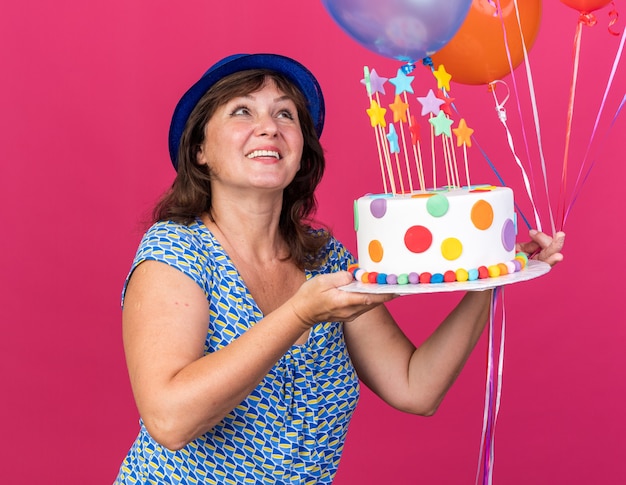 Happy middle age woman in party hat with colorful balloons holding birthday cake looking up with smile on face celebrating birthday party standing over pink wall